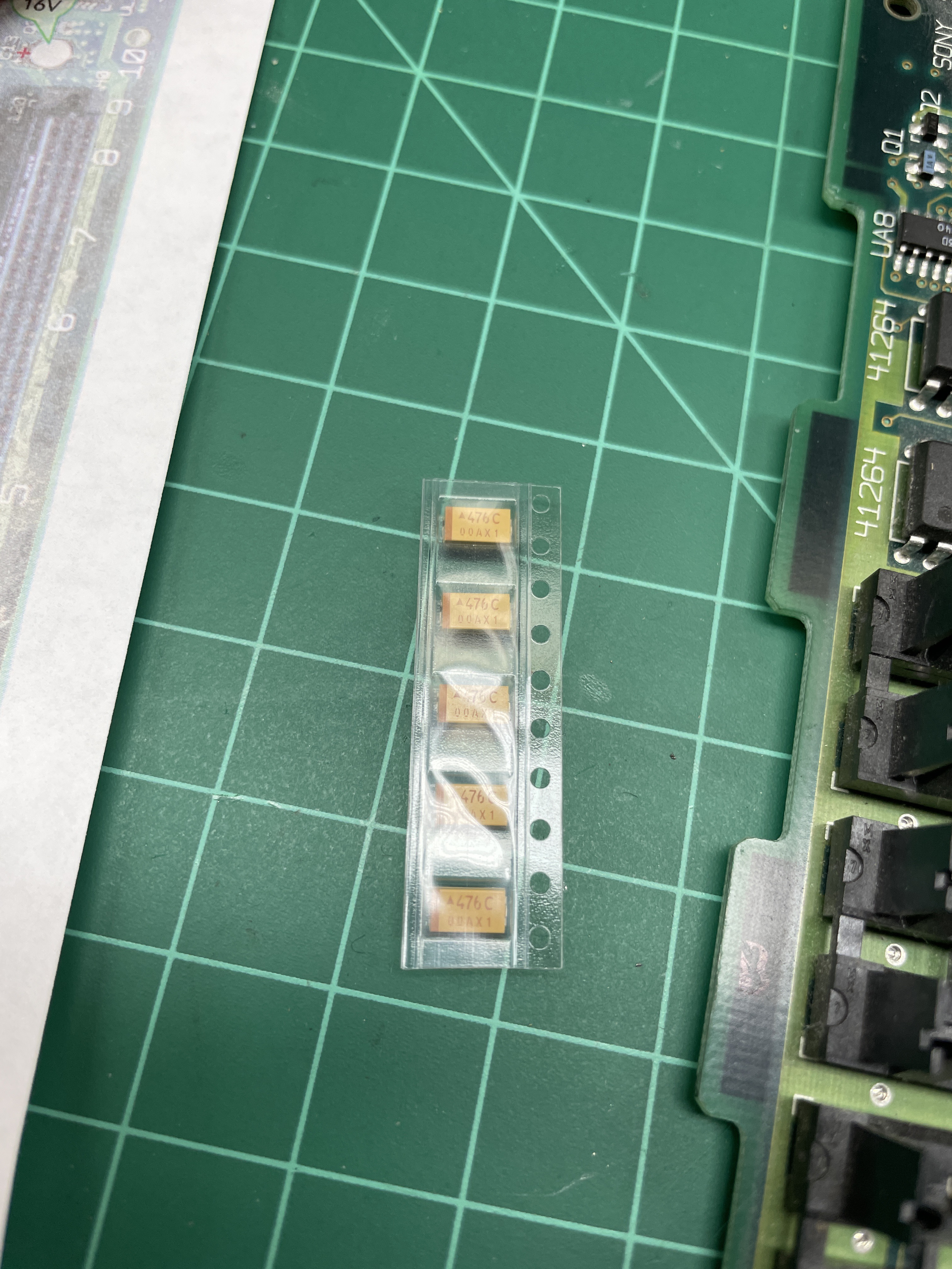 A strip of new surface-mount tantalum capacitors in their shipping tape, sitting on a green cutting mat next to the logic board