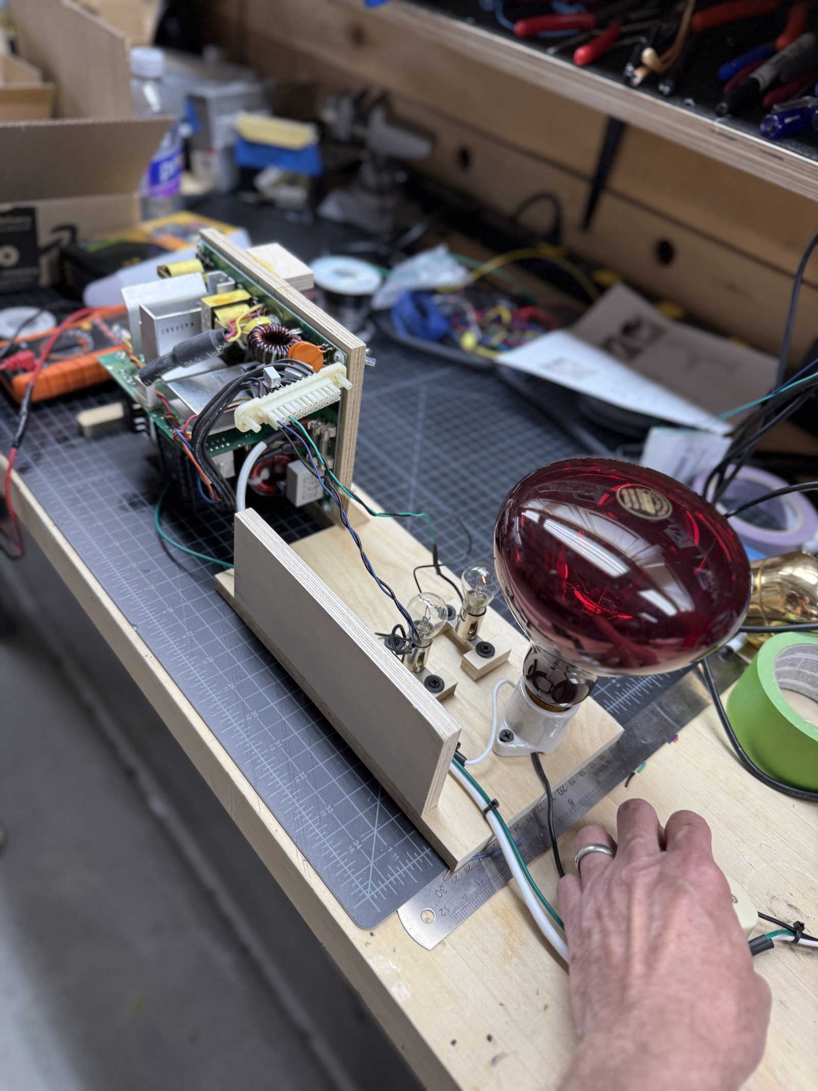 Plywood bench jig holding the PEC4044B main board upright, with 1156 bulb dummy loads and the 250W heat lamp ballast mounted alongside