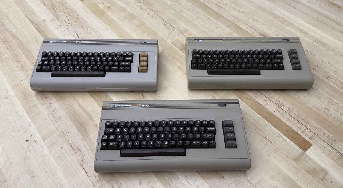 Three Commodore 64s arranged on a hardwood floor: the new FPGA-based Commodore 64 Ultimate at upper left, the original 1980s breadbin at lower middle, and TheC64 from Retro Games Ltd. at upper right